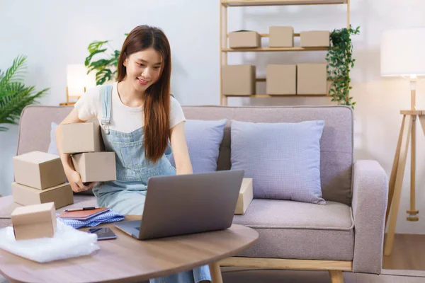 Online seller concept, Female entrepreneur holds parcel boxes and checks online orders on laptop.