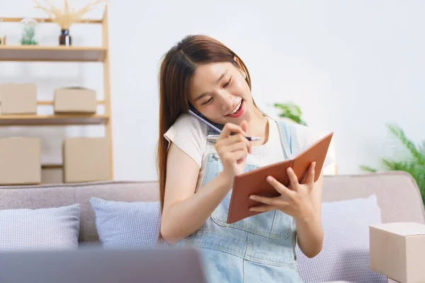 Online merchant concept, Female entrepreneur taking notes in notebook while talking on smartphone.