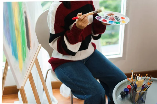 The art concept, Asian female artist in sweater holding paint brush and color palette in art studio.