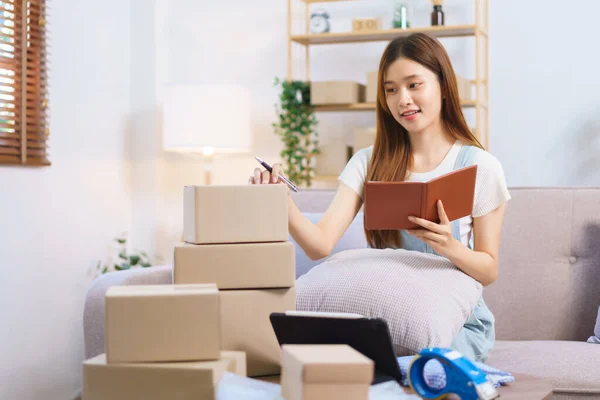 Online merchant concept, Female entrepreneur writes on parcel box after reading address on notebook.