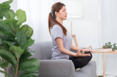 Yoga exercise concept, Young Asian woman doing yoga exercise in lotus pose on couch in living room.