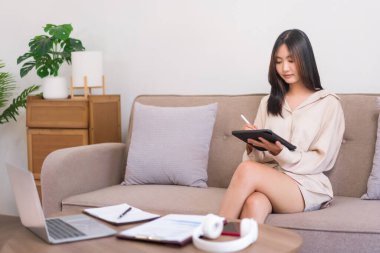 Evde rahatlama kavramı, genç Asyalı kadın kanepede otururken çalışmak için tablete yazı yazıyor..