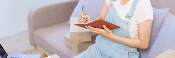 Online merchant concept, Female entrepreneur taking notes in notebook while talking on smartphone.