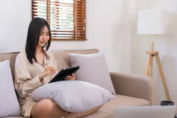 Concept of activity in living room, Asian woman writing on tablet to working while sitting on sofa.