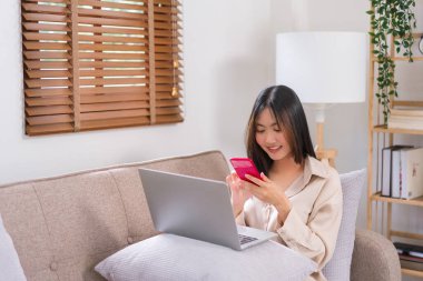 Evde rahatlama kavramı, genç Asyalı kadın kanepede arkadaşlarıyla sohbet etmek için akıllı telefonu kullanır..