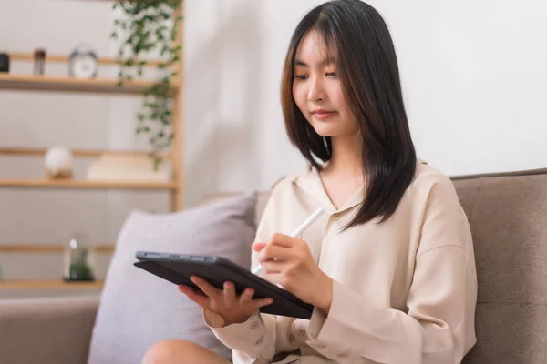 Concept of activity in living room, Asian woman writing on tablet to working while sitting on sofa.
