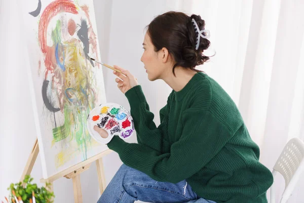 Creative of art concept, Young asian woman hold paintbrush and color palette while looking artwork.
