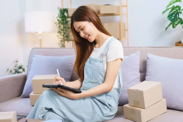 Online merchant concept, Female entrepreneur checks online orders on tablet to prepare parcel boxes.