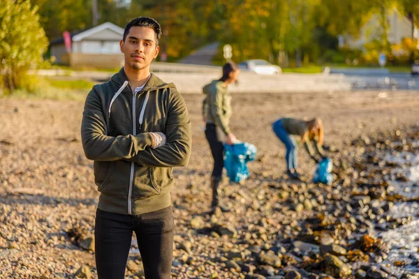 Serieuze man met zijn armen gekruist op het strand met een groep vrijwilligers op zonnige dag — Stockfoto