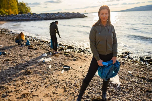 Smiling young woman cleaning beach for plastic with volunteers during beautiful sunset — Stockfoto