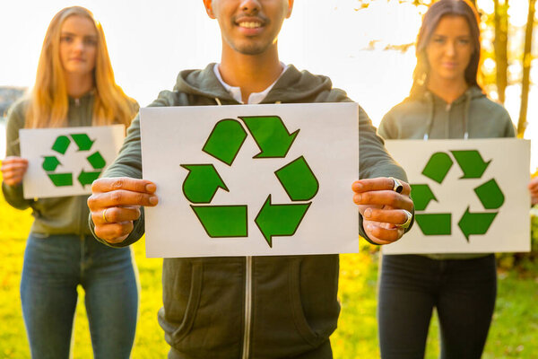Team of young volunteers holding recycling symbol placards. Portrait of smiling man and women standing at park. They are protecting environment.