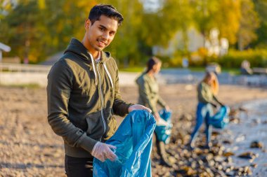 Full length portrait of young man picking up garbage in bag. Multi-ethnic group of volunteers are cleaning beach against sky. They are protecting environmental from damage.