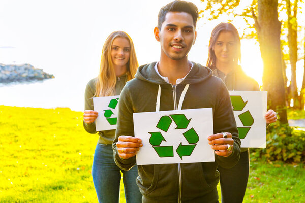 Team of young volunteers holding recycling symbol placards. Portrait of smiling man and women standing at park. They are protecting environment.