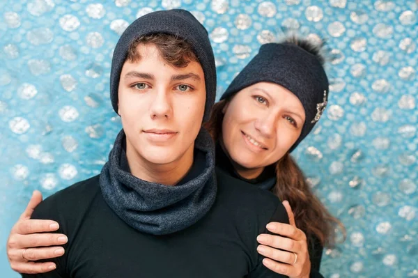 Mom hugs a beautiful teenage son on a blue background. Son and Mom are dressed in warm winter hats and scarves. They are happy that they are well prepared for winter and will not freeze in severe frost