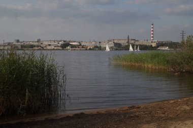 Tam renkli yatay fotoğraf. Endüstriyel yaz manzarası. 18. yüzyılda bir fabrika barajının oluşturduğu bir gölet. Ön planda sazlıklar var. Arka planda, fabrika binalarının arka planına karşı bir yelkenli yarışı var..