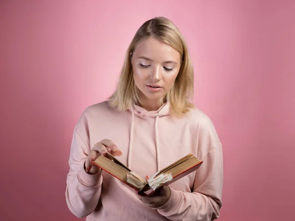 Очаровательная молодая студентка с книгой в руках. Молодая милая блондинка в розовой толстовке — стоковое фото