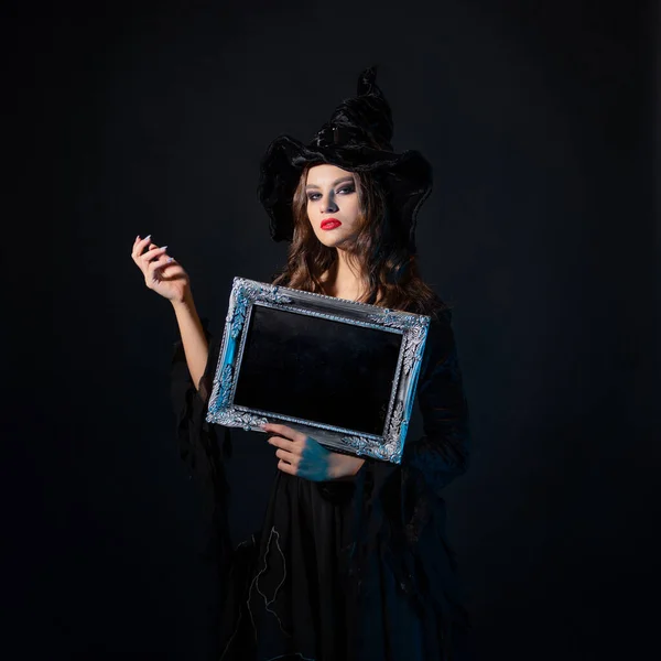 Une jeune belle sorcière au chapeau pointu tient dans ses mains un cadre argenté avec un endroit noir pour le texte, — Photo