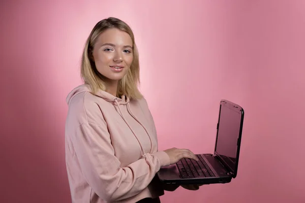 young woman with a laptop, a student or a freelancer. A charming blonde in a pink hoodie