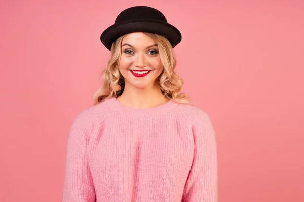 Μια νέα, ελκυστική γυναίκα με ένα ζεστό ροζ πουλόβερ και ένα μαύρο καπέλο σε ροζ φόντο., — Φωτογραφία Αρχείου