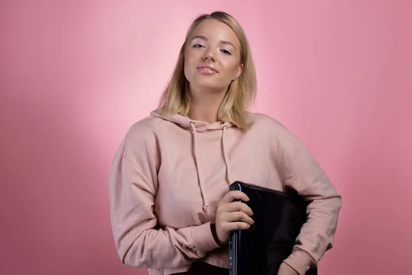 Fiatal nő laptoppal, tanulóval vagy szabadúszó. Egy bájos szőke, rózsaszín kapucnis pulcsiban. — Stock Fotó