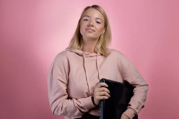 Giovane donna con un computer portatile, uno studente o un libero professionista. Un'affascinante bionda con una felpa rosa — Foto Stock