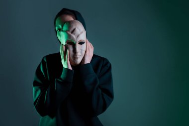 Maskenin arkasına saklanan koyu renk kapüşonlu genç bir kadın yüzünü maskeyle gizliyor.,