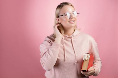 Pembe kapüşonlu ve gözlüklü genç mutlu bir öğrenci elinde bir kitap tutuyor.,