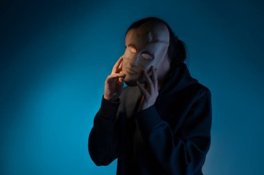 Maskenin arkasına saklanan koyu renk kapüşonlu genç bir kadın yüzünü maskeyle gizliyor.,