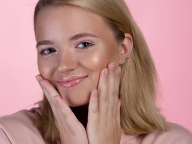 Pembe kapüşonlu tatlı sarışın mutlu bir şekilde gülümseyip eliyle yüzüne dokunuyor..