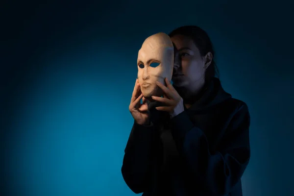 Hinter einer Maske versteckt sich eine junge Frau in dunklem Kapuzenpulli, die ihr Gesicht mit einer Maske verdeckt, — Stockfoto