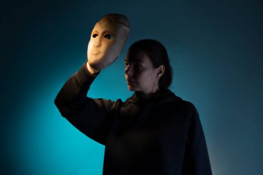 Maskenin arkasına saklanan koyu renk kapüşonlu genç bir kadın yüzünü maskeyle gizliyor.,