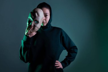 Maskenin arkasına saklanan koyu renk kapüşonlu genç bir kadın yüzünü maskeyle gizliyor.,