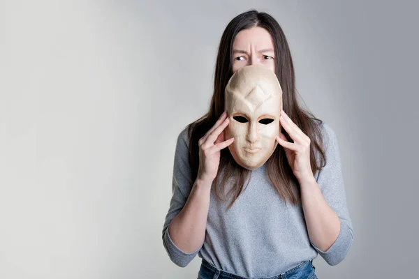 Młoda kobieta trzyma maskę w dłoniach, ukrywając połowę twarzy, przerażone spojrzenie na maskę.. — Zdjęcie stockowe