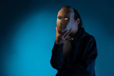 Maskenin arkasına saklanan koyu renk kapüşonlu genç bir kadın yüzünü maskeyle gizliyor.,