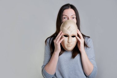 Genç bir kadın elinde maske tutuyor, yüzünü yarım yamalak gizliyor, maskenin üzerinden şaşırmış bir bakış..