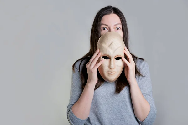 O femeie tânără ține o mască în mâini, ascunzându-și fața pe jumătate, o privire surprinsă peste mască. — Fotografie, imagine de stoc
