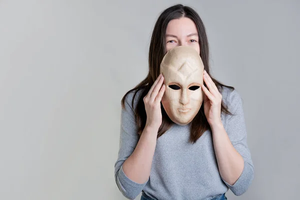 O femeie tânără ține o mască în mâini, ascunzându-și fața pe jumătate, o privire vicleană peste mască. — Fotografie, imagine de stoc