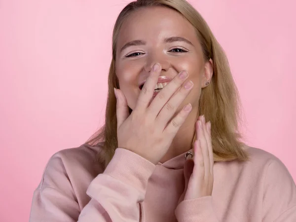 분홍색 후드를 쓴 귀여운 젊은 금발 소녀가 그녀의 입 과 얼굴을 그녀의 손으로 즐겁게 가리고 웃는다. — 스톡 사진