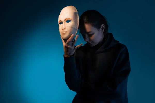 Chowając się za maską, młoda kobieta w ciemnej bluzie ukrywa twarz w masce., Zdjęcia Stockowe bez tantiem
