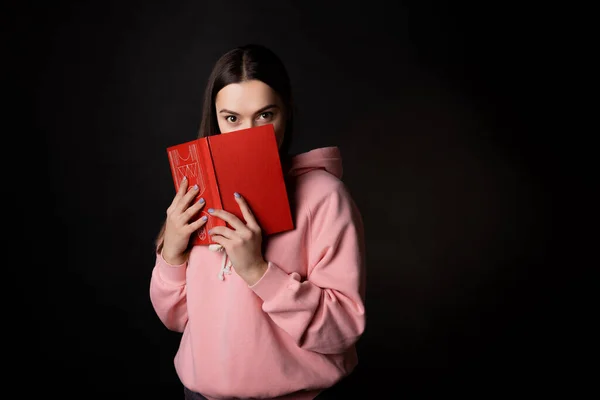 A young cute brunette in a pink hoodie, reading a book. A student with an open book in her hands, — Fotografie, imagine de stoc