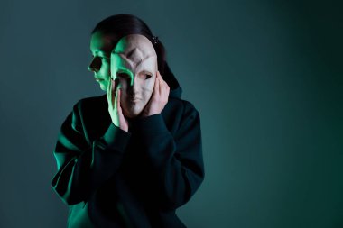 Maskenin arkasına saklanan koyu renk kapüşonlu genç bir kadın yüzünü maskeyle gizliyor.,