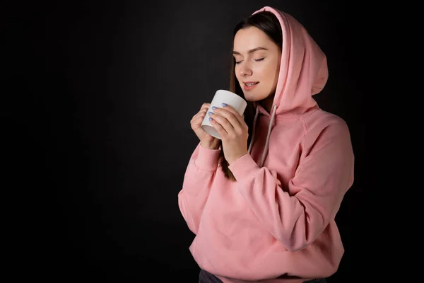 Cute young brunette in a pink hoodie drinking tea or coffee from a large mug, — Foto Stock