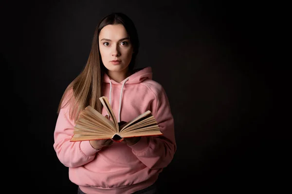 Mladá roztomilá brunetka v růžové mikině, čte si knížku. Studentka s otevřenou knihou v rukou, — Stock fotografie