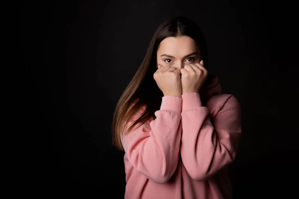 A cute young brunette in a pink hoodie hides her face with her hands, — Stock Photo, Image