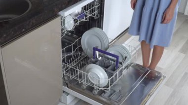 Ev hanımı bulaşık makinesinden temiz tabaklar ve kupalarla üst rafı itiyor.