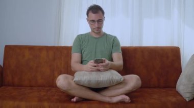Lotus pozisyonunda telefonu olan bir adam kanepeye oturur ve akıllı telefondan cevap verir. Yüksek kalite 4k görüntü
