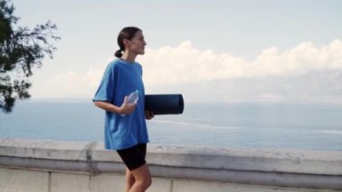 Elinde yoga minderi ve su olan bir kadın antrenman için deniz boyunca yürür. Yüksek kalite 4k görüntü