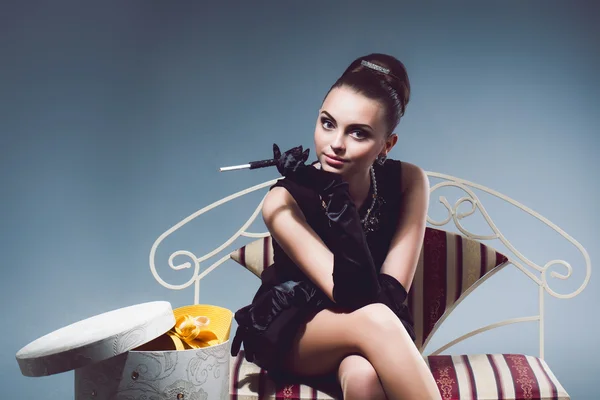 Young luxurious rich girl in black dress sitting on a sofa holding cigarette in hand with long gloves and jewelry or ornaments, next to the box for hats in the studio — Stock Photo, Image