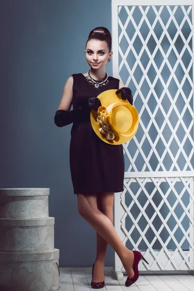 Chica joven en un pequeño vestido negro corto sosteniendo un sombrero amarillo en la mano, guantes negros largos, cajas de sombrero en el estudio. Sorprendida dama de lujo.Imagen de una chica en retro — Foto de Stock