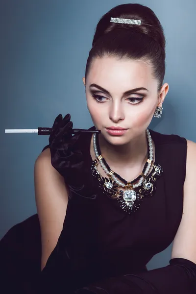 Portrait young girl in a black dress looks down thoughtfully keep the cigarette holder with cigarette in hand with long gloves and jewelry  on the neck, — Stock Photo, Image